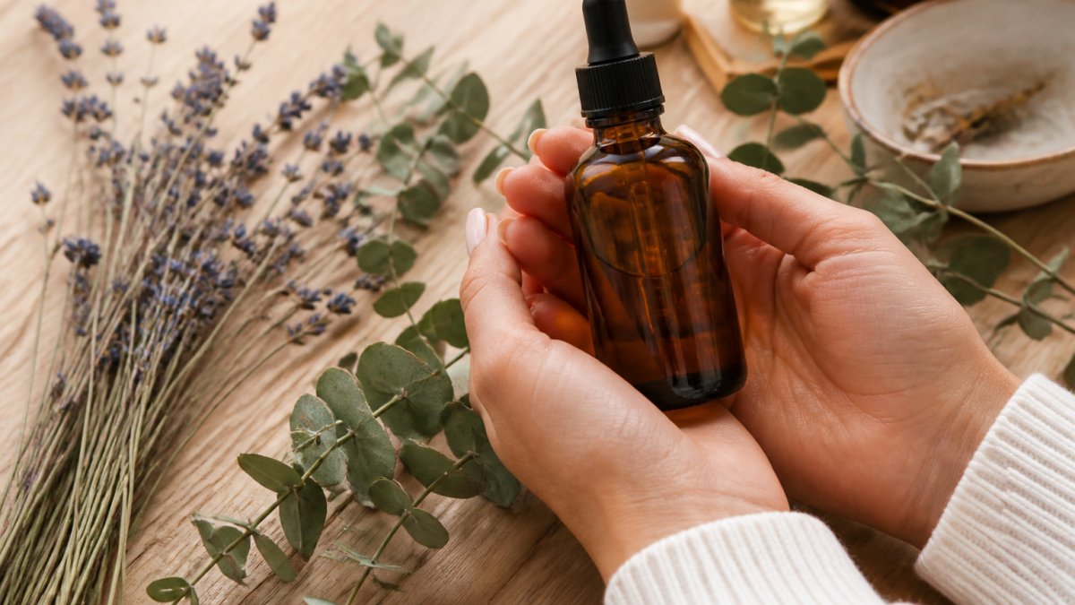 Close-up of hands holding a small amber glass bottle of body oil with dried lavender and eucalyptus sprigs on a light wooden table, soft natural light