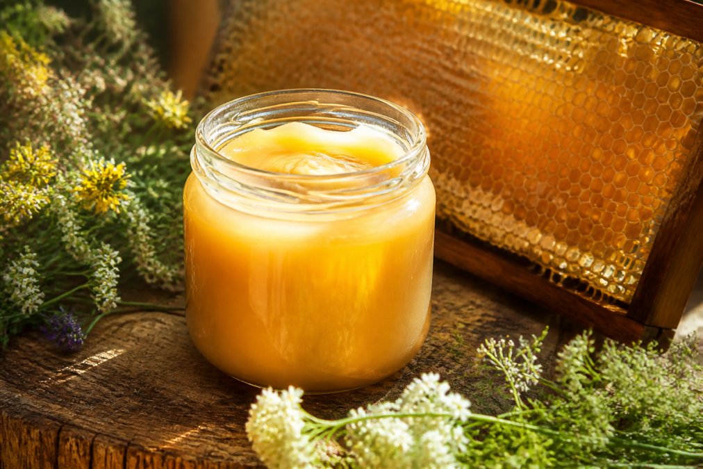 Nahaufnahme eines geöffneten Glases cremigen Sommerblütenhonigs mit goldener Farbe, Honigwabe im Hintergrund, Sonnenlicht scheint durch das Glas und wirft warme Reflexionen auf die Oberfläche