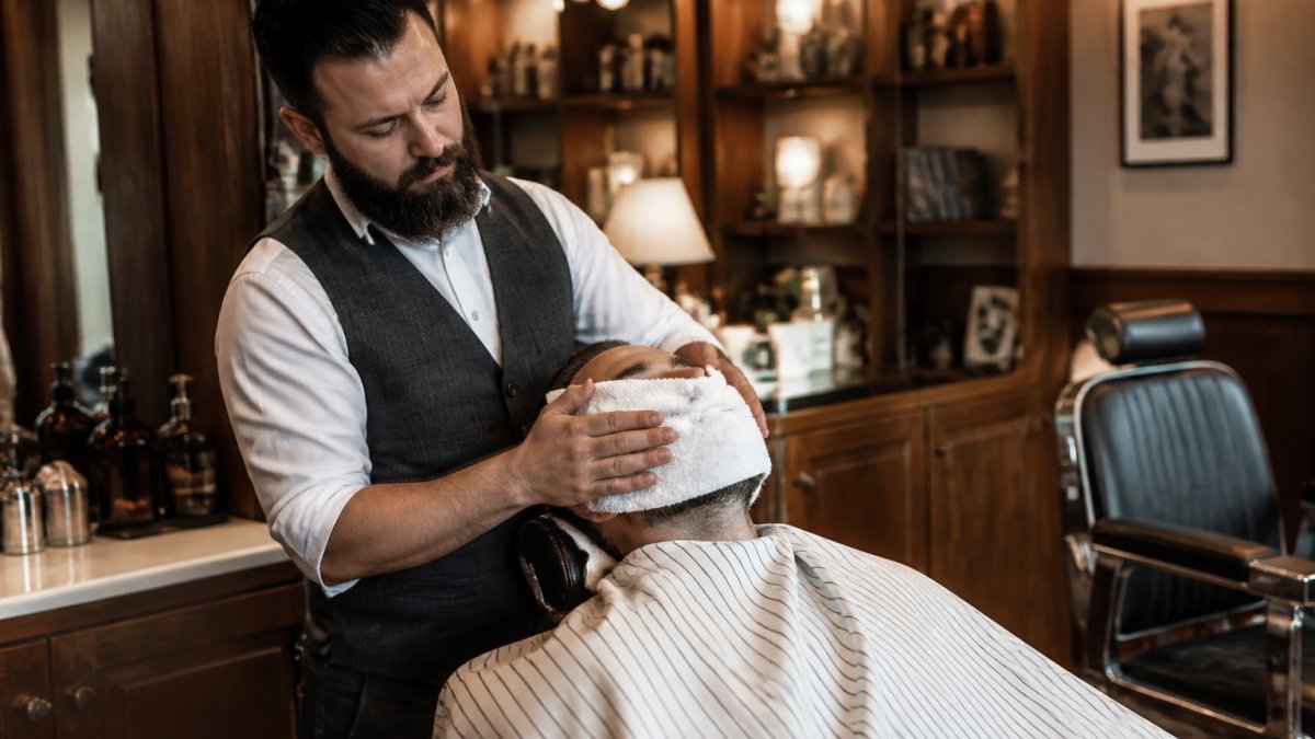 Professional barber applying a steaming hot towel to client's face in upscale barbershop setting with warm lighting and leather chairs, preparing for a straight-razor shave
