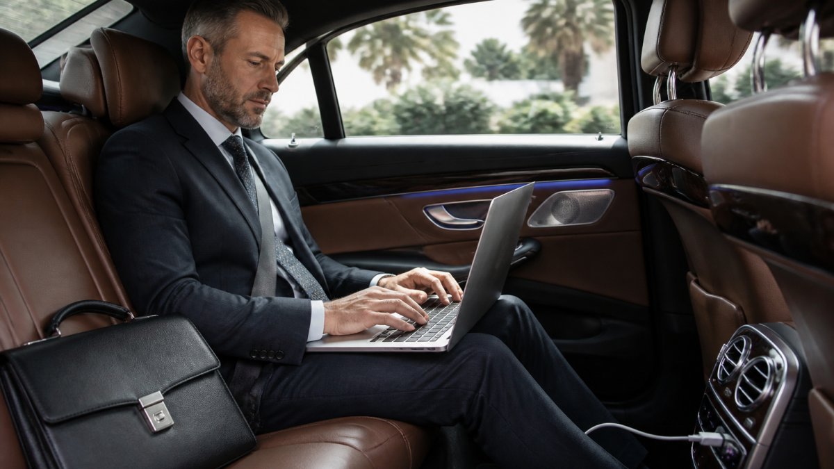 Executive working on laptop in luxury sedan rear seat with ambient lighting, charging ports visible, professional briefcase on adjacent seat, natural window light
