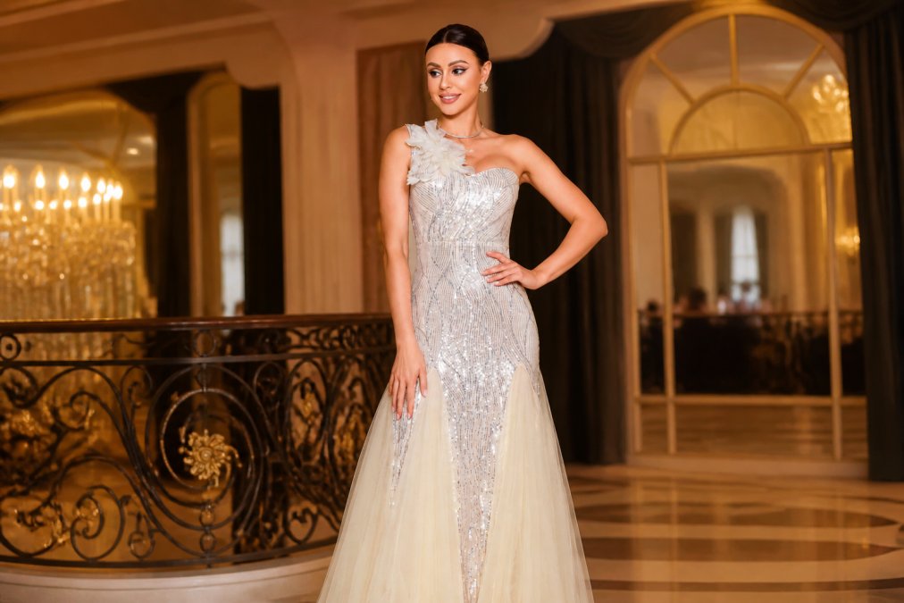 Woman wearing an elegant sequined cocktail dress in rich jewel tones, paired with strappy metallic heels and minimalist drop earrings, standing in a softly lit ballroom setting