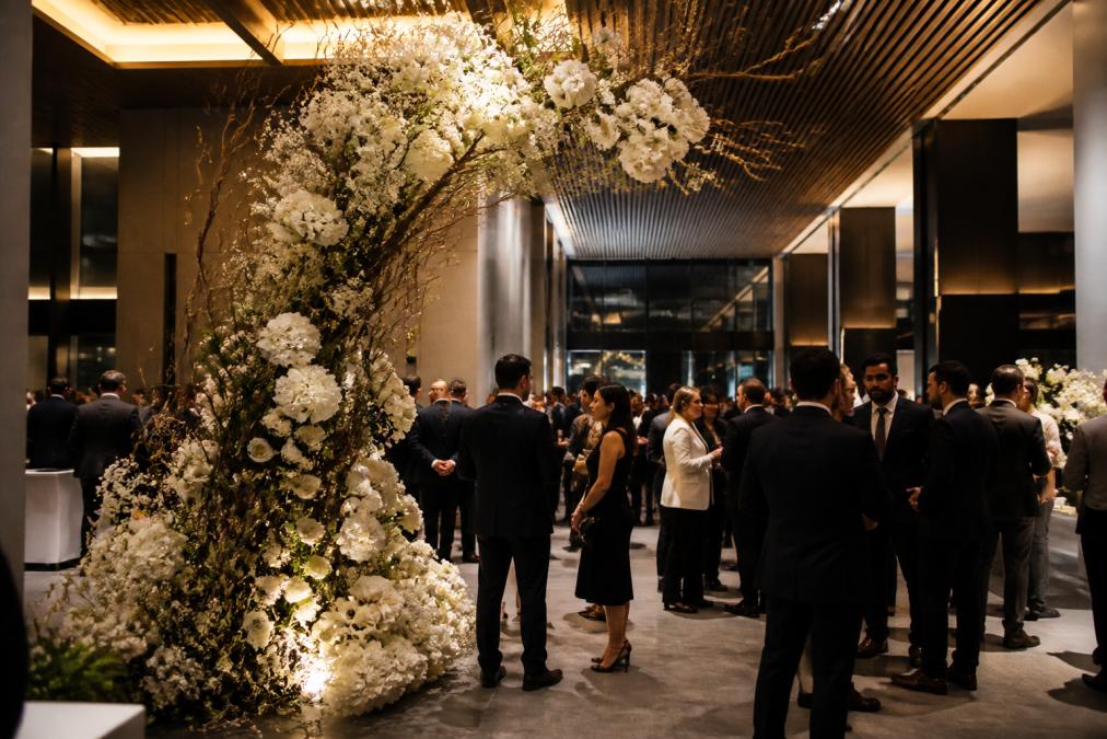 A modern corporate event space with a large sculptural floral installation in white and cream blooms, elegantly dressed attendees mingling beneath warm architectural lighting