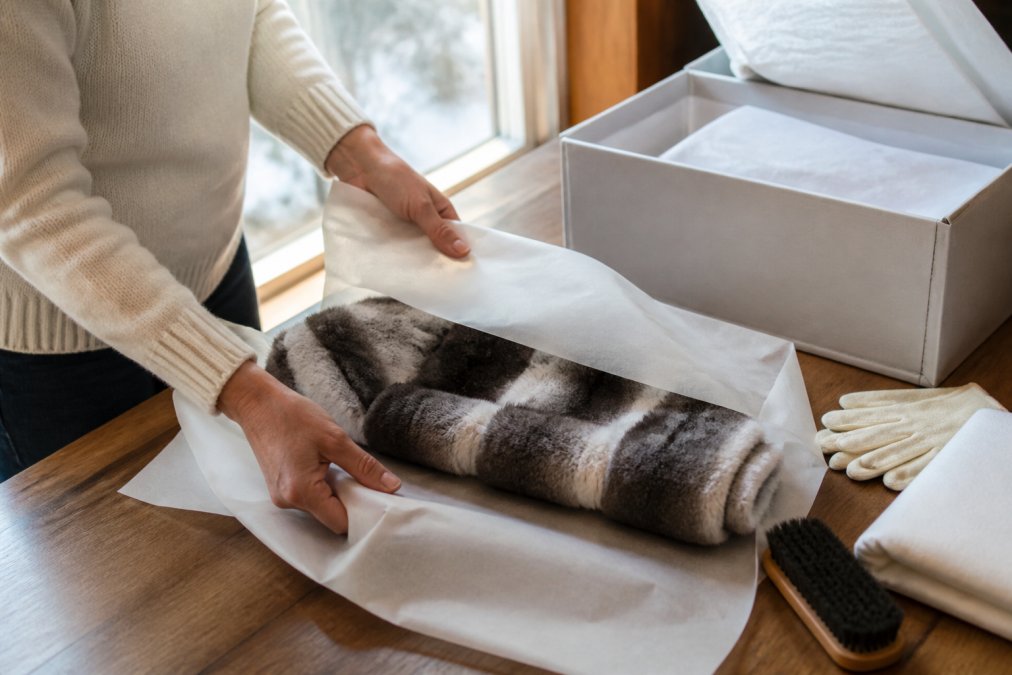 Hands carefully folding a luxurious chinchilla wrap in soft acid-free tissue paper, with storage boxes and preservation materials visible on a wooden table in natural daylight