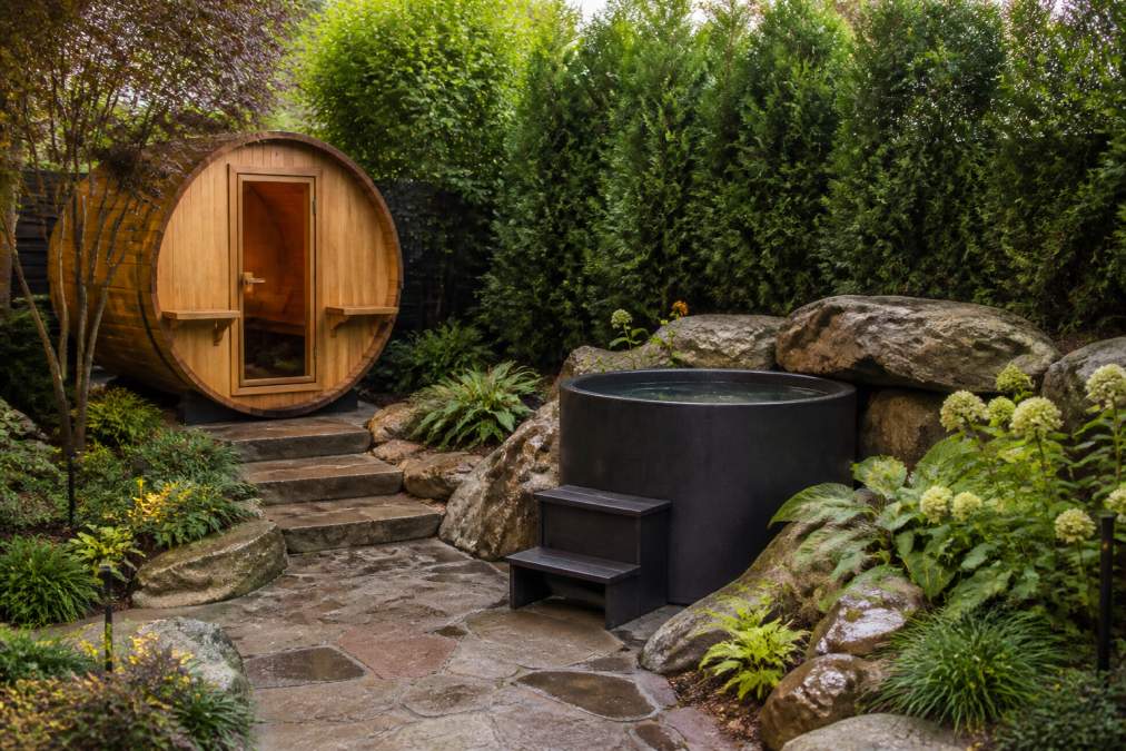 Luxury sauna and cold plunge positioned in private garden corner with tall privacy screening plantings, natural stone hardscaping, and softscape plantings creating complete wellness retreat sanctuary