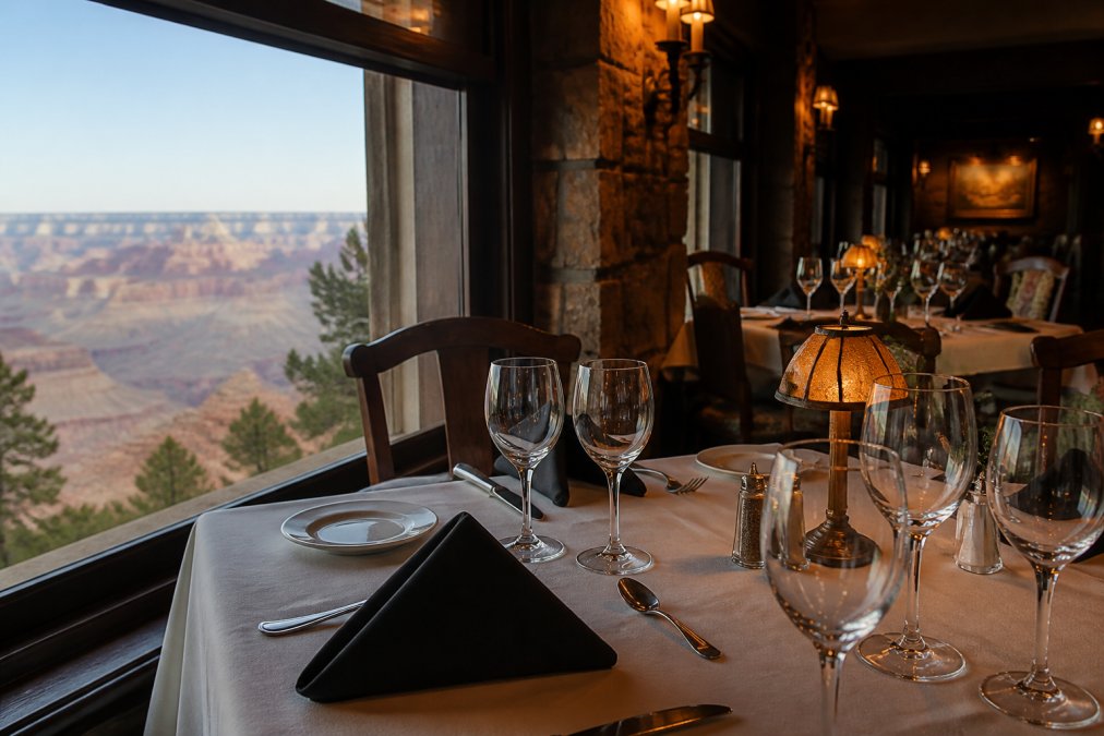 An elegant table setting inside El Tovar's historic stone-and-dark-wood dining room, white linen tablecloths, polished wine glasses catching warm ambient light, with a window framing the Grand Canyon rim in the background