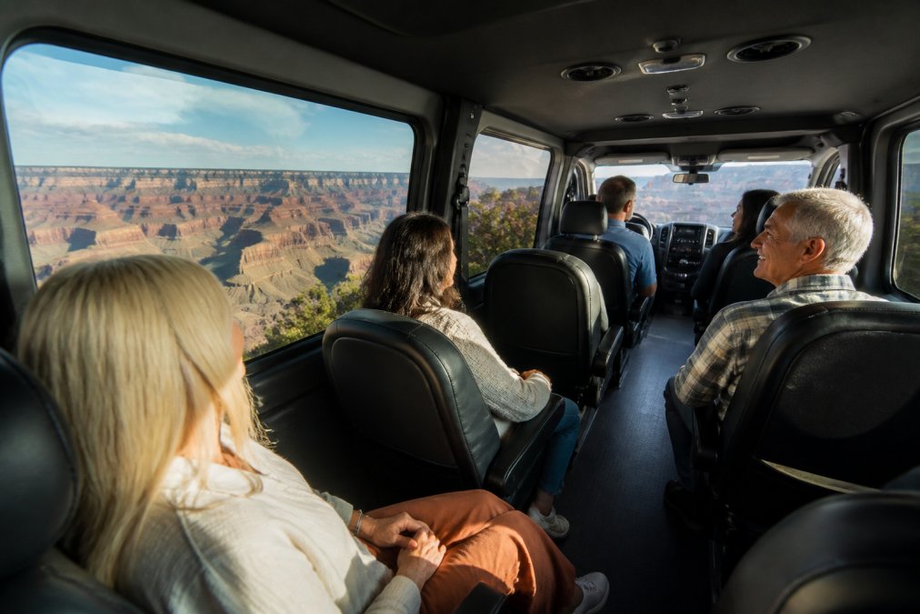 Passengers enjoying panoramic views of the Grand Canyon through large windows of a comfortable Ford Transit van interior, with flexible seating and natural lighting showing the canyon landscape