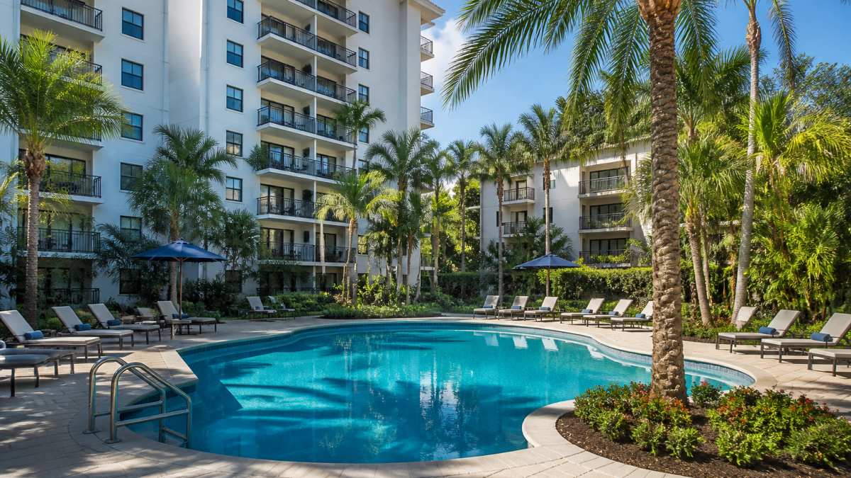 Lush landscaped grounds with mature palm trees, manicured gardens, and a sparkling resort-style pool surrounded by comfortable lounge seating on a sunny afternoon