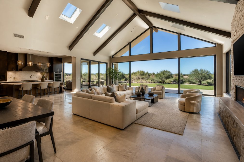 Luxury home interior with vaulted ceilings and skylights bathing the space in golden natural light, wide-angle view showcasing an open-concept living area with high-end furnishings, architectural details, and floor-to-ceiling windows overlooking a manicured landscape