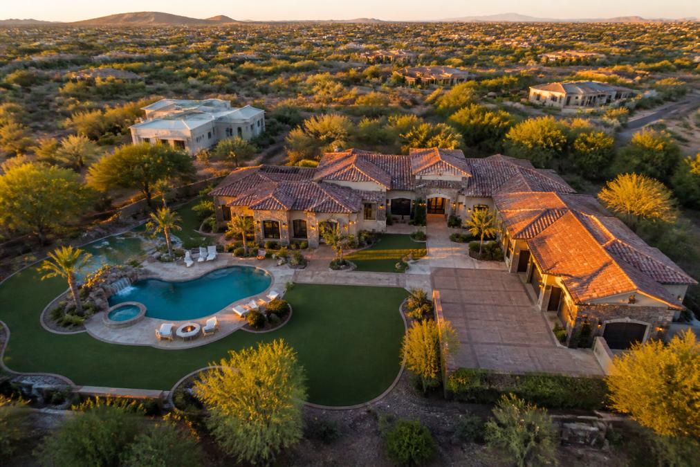 Aerial view of a luxury estate at golden hour, drone shot capturing the entire property including manicured grounds, resort-style pool with waterfall feature, multi-car garage, and surrounding landscape, showing property scale and architectural design from above