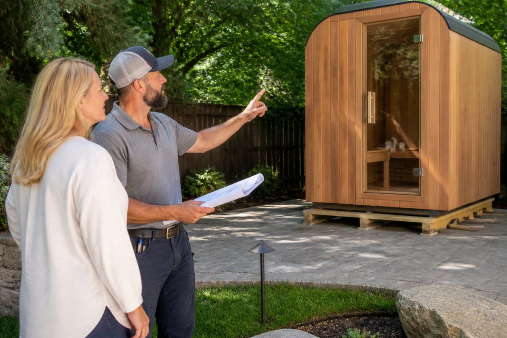 A professional installer in clean work clothes reviewing sauna placement plans with a homeowner in a bright backyard, pointing at a cedar sauna cabin being positioned on a stone patio