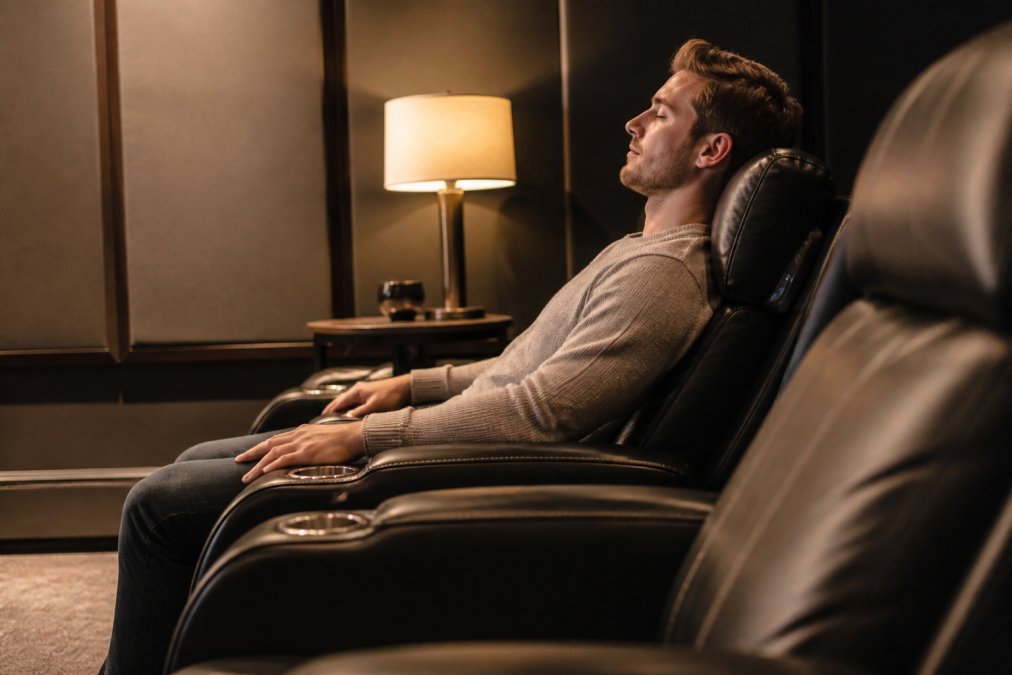 Person seated in luxury home theater recliner demonstrating proper lumbar support alignment, with backrest cradling spine and shoulders relaxed against headrest, warm ambient lighting from side table