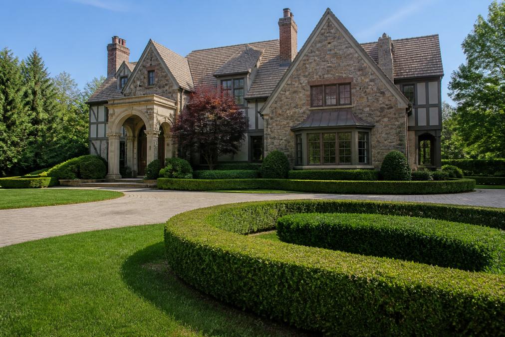 Elegant stone and timber French Normandy Tudor mansion with manicured hedges, circular driveway, and ornate entrance columns in soft afternoon light