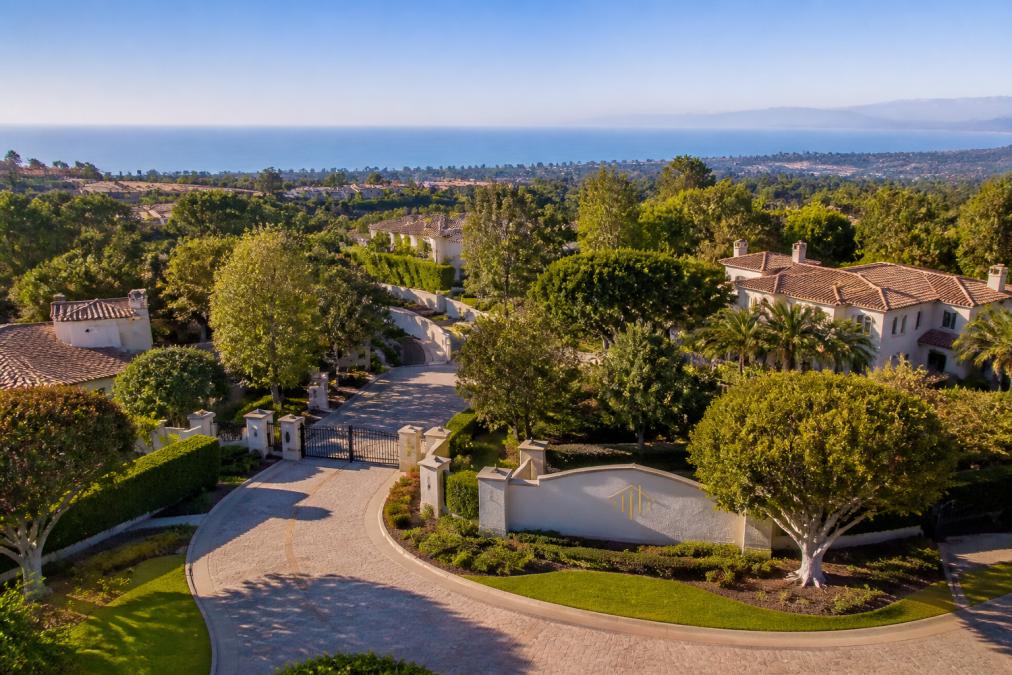 Aerial view of a gated estate community with manicured grounds, tree-lined streets, winding driveways, and glimpses of ocean views in the distance under clear morning light