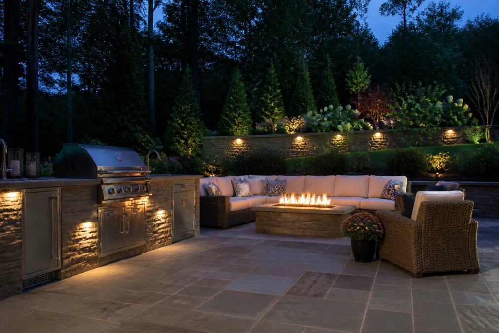 An elegant outdoor living area in a Scarsdale backyard at dusk, featuring a stone patio, an outdoor kitchen with a grill, and a comfortable seating area with a fire pit