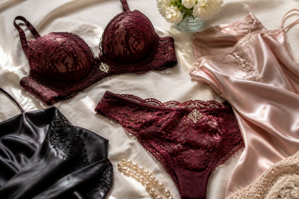 Close-up of luxurious silk and lace lingerie items arranged elegantly on a soft cream linen surface, showcasing delicate embroidery, rhinestone accents, and sumptuous fabric textures in natural window light