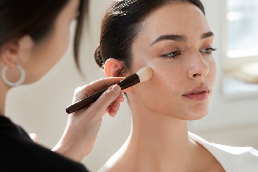 Close-up of a makeup artist's hands using a soft handcrafted brush to apply foundation to a model's cheek, focusing on the radiant skin-like finish, natural window light, neutral tones, shallow depth of field