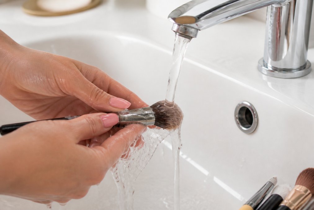 Close-up of hands gently cleaning makeup brushes under running water with a mild cleanser, showing proper technique and care with natural lighting