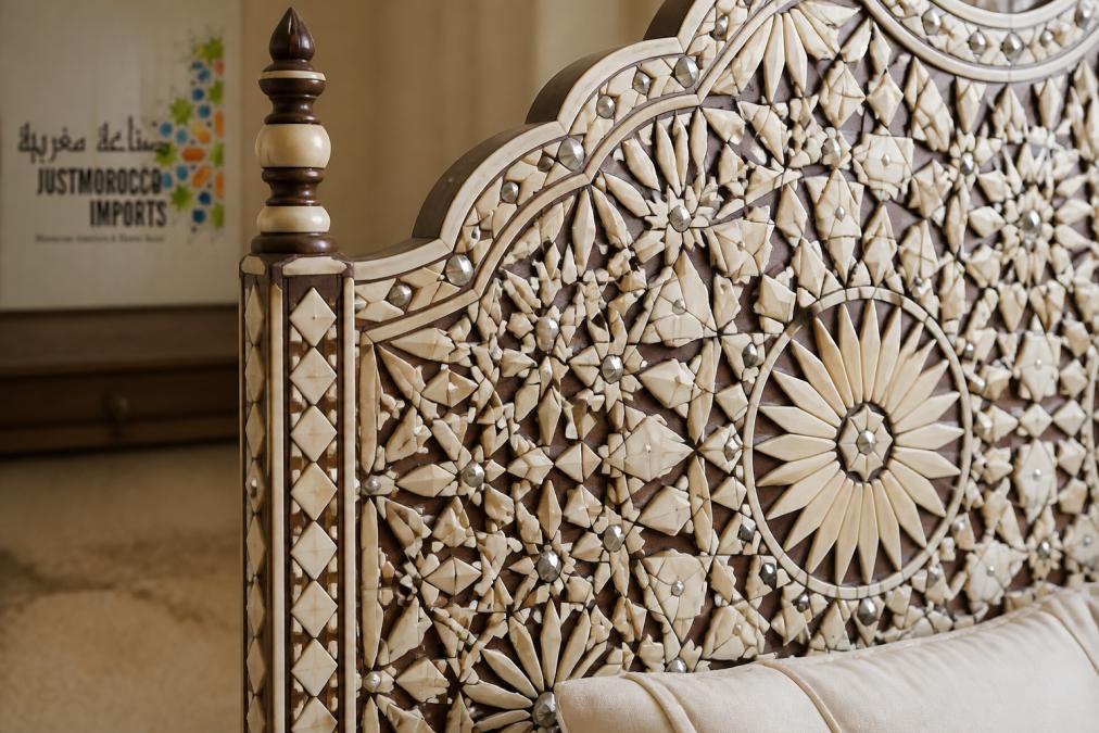 Close-up of intricate camel bone inlay details on a wooden bed headboard, showing hand-chiseled geometric patterns and silver metal accents with warm natural light highlighting the texture and dimensional carving