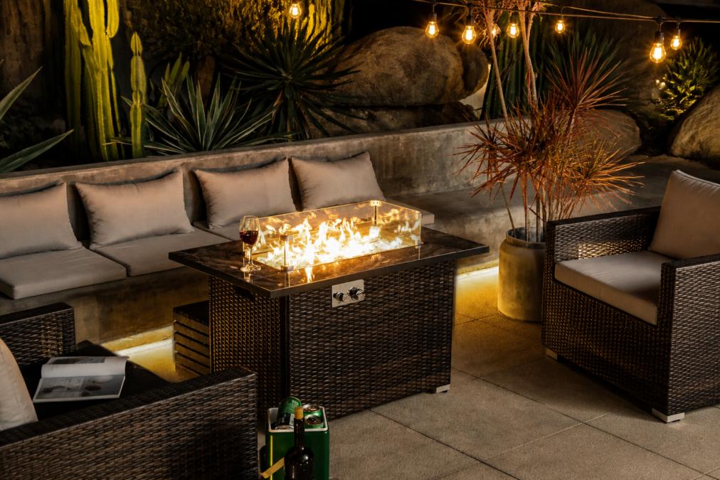 Elegantly styled outdoor patio at dusk with a marble-topped fire table as the centerpiece, surrounded by comfortable seating with neutral cushions, warm ambient lighting from string lights overhead, and coordinated outdoor furniture creating a sophisticated entertaining space