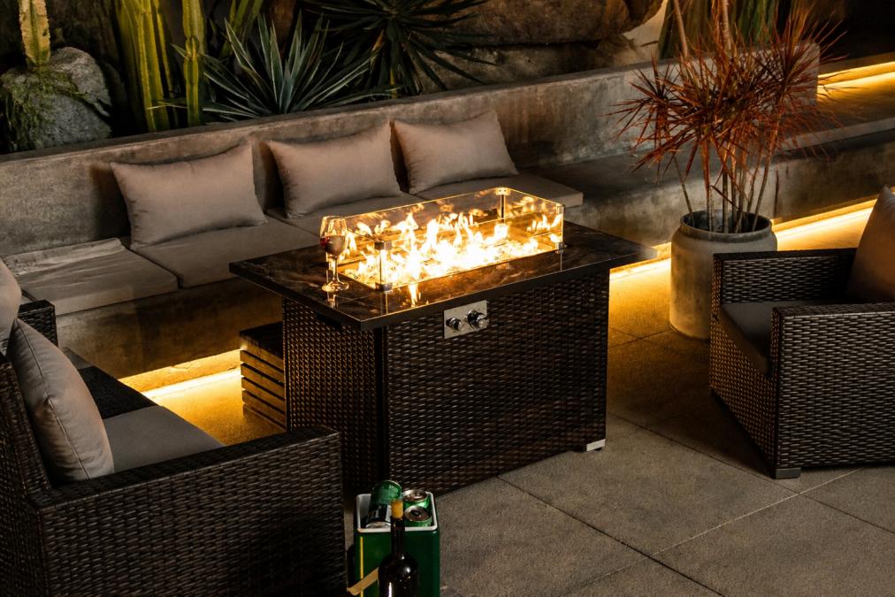 Luxury backyard landscape featuring a modern fire pit table integrated into hardscaping with stone pavers in geometric patterns, decorative plantings of ornamental grasses and low shrubs, and architectural elements creating defined zones that establish an outdoor retreat atmosphere