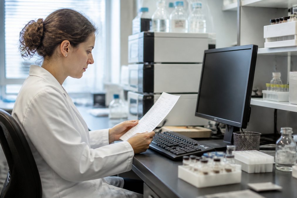 Research scientist at a modern laboratory workstation carefully reviewing supplier documentation and quality reports on a computer screen, with peptide vials and analytical equipment visible in the soft background lighting