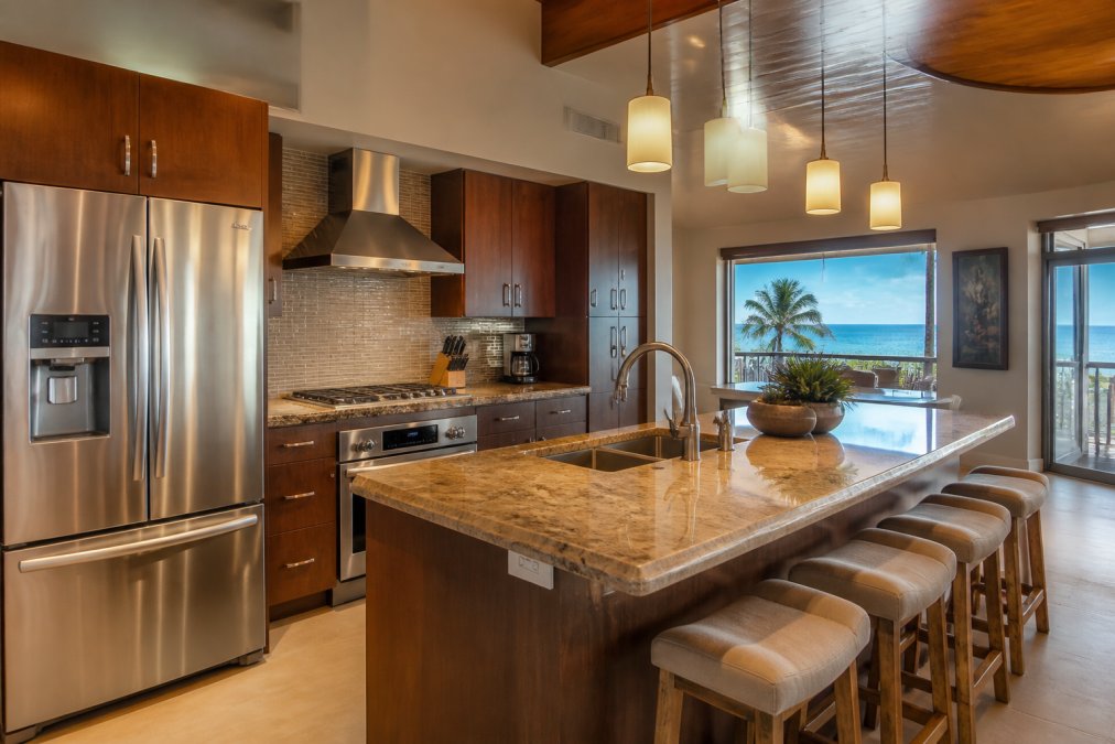 A fully equipped modern luxury kitchen inside a vacation rental condo, with high-end stainless steel appliances, stone countertops, pendant lighting, and a window framing a view toward the Pacific Ocean in afternoon light