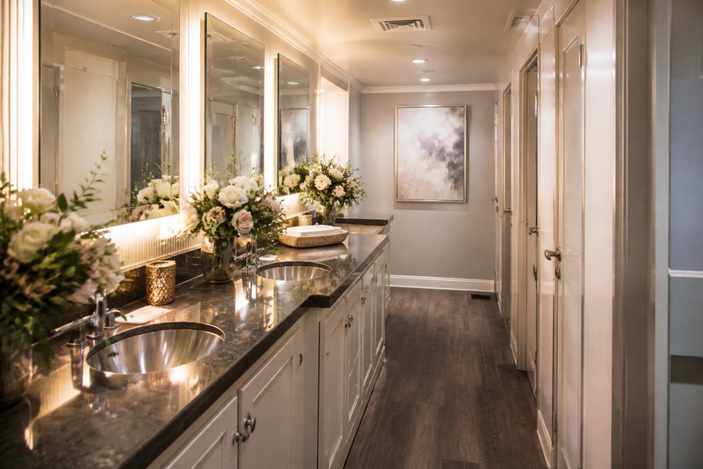 Interior of a luxury restroom trailer decorated for a wedding, showing vanity mirrors with warm lighting, fresh flowers on the counter, polished chrome fixtures, and a climate-controlled space with hardwood-style flooring and soft overhead lighting