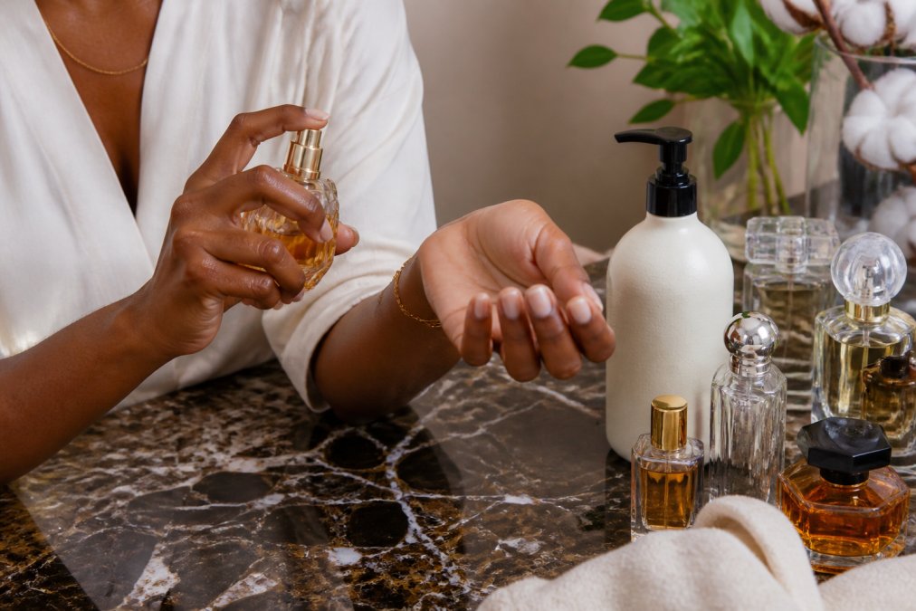 Close-up of hands applying fragrance to pulse points on wrist and neck, with luxury perfume bottles and unscented base lotion on a marble surface