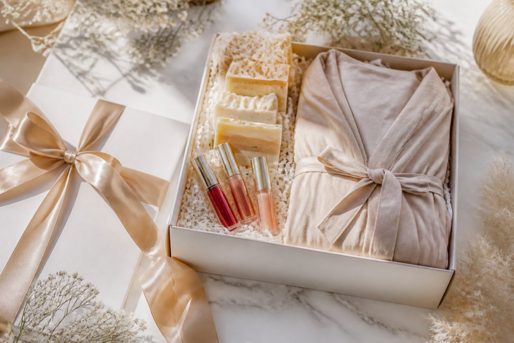 Elegantly wrapped luxury gift set with premium handcrafted soaps, tinted lip oils, and bamboo loungewear artfully displayed on a white marble surface with soft natural morning light highlighting textures and packaging