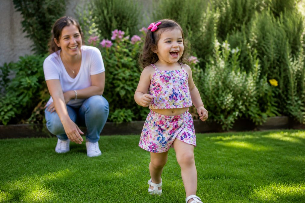 A happy toddler girl wearing a colorful floral skort set playing in a sunlit backyard garden, with a parent nearby smiling, natural summer light, candid lifestyle photography