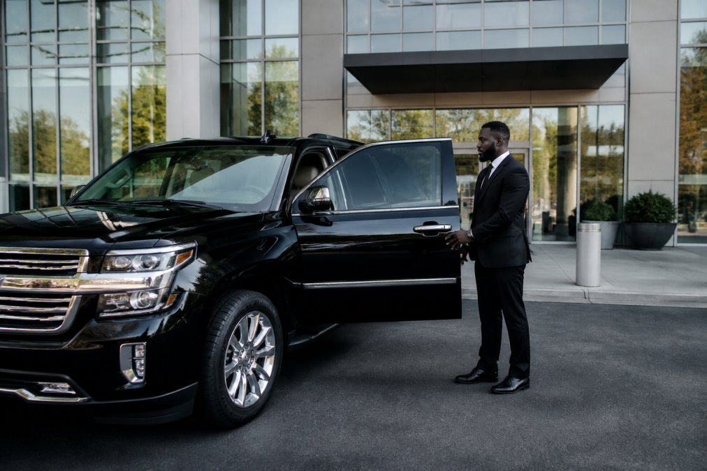 Sleek black luxury SUV parked outside a modern glass office building with professional chauffeur in dark suit standing beside open passenger door, morning light reflecting off polished exterior