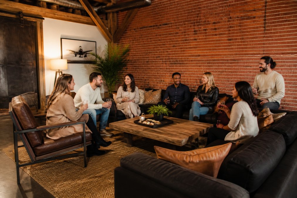 A group of eight people gathered in a spacious, high-end living room with exposed brick, vintage timber beams, and modern furnishings, enjoying drinks and conversation