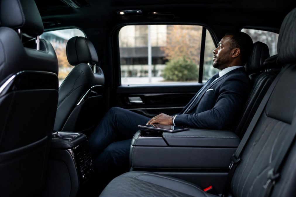 A business executive in a tailored suit relaxing in the spacious rear seat of a black luxury SUV, with a tablet on the fold-down armrest and soft ambient lighting visible in the cabin