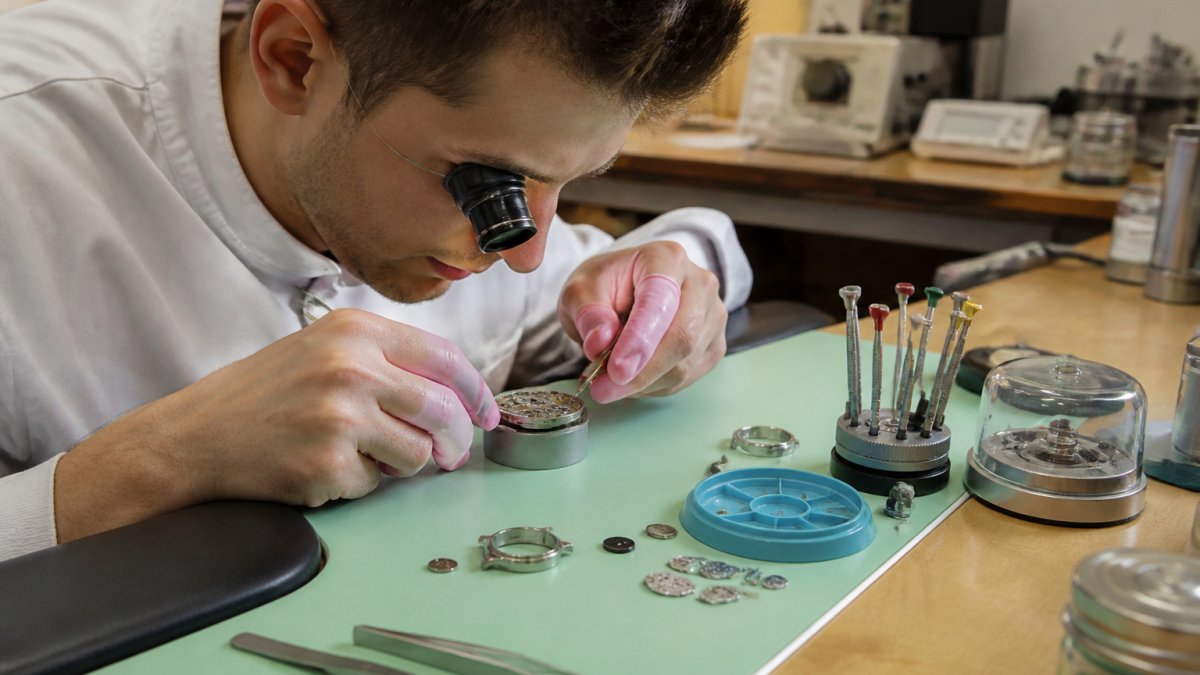 Professional watchmaker inspecting luxury watch movement under magnification lamp with precision tools and components visible on workbench