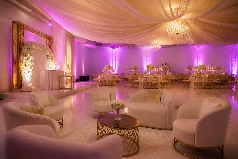 An elegant wedding reception setup inside a luxury venue with draped ceiling fabric, up-lighting, and lounge furniture arranged for guests