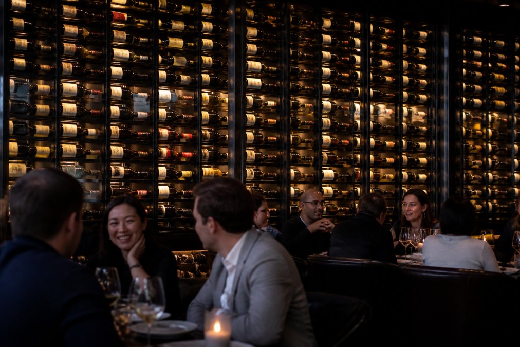 Upscale restaurant dining room with dramatic floor-to-ceiling matte black wine wall display featuring bottles in label-forward orientation, warm ambient lighting illuminating the collection, and diners in foreground enjoying the sophisticated setting