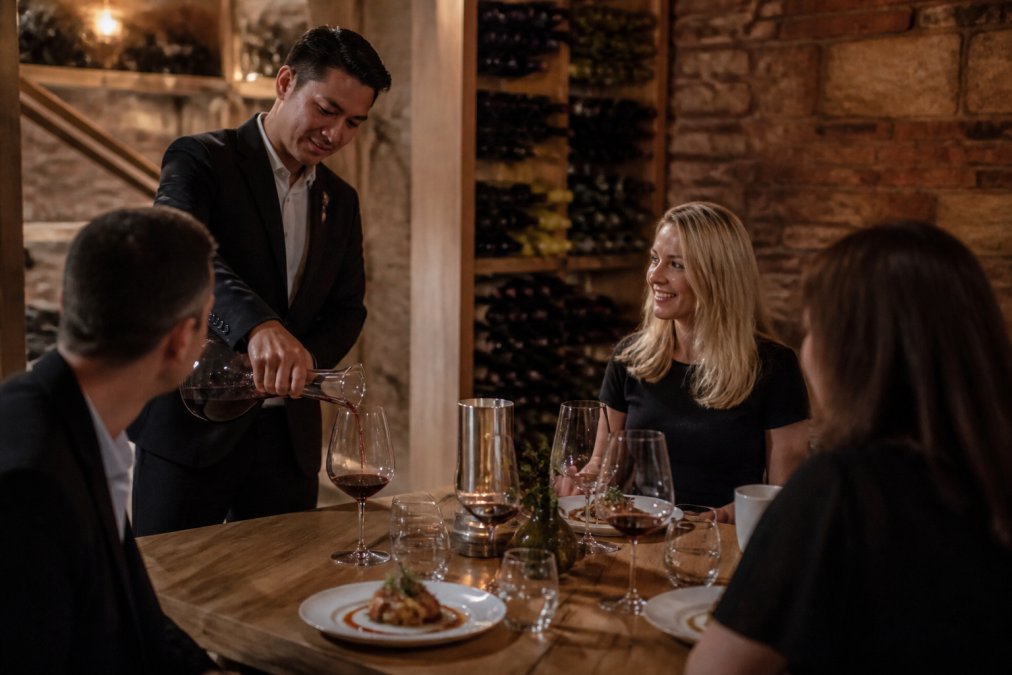 Professional sommelier pouring premium wine into crystal glass beside elegantly plated gourmet dish featuring local regional produce, natural vineyard cellar lighting
