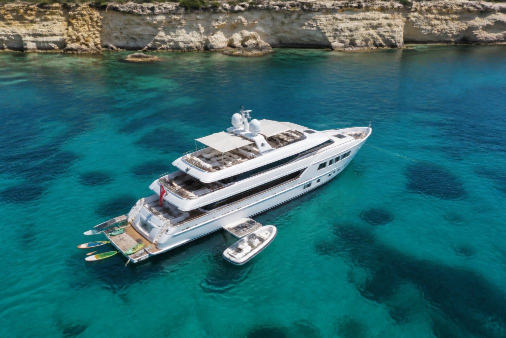 Aerial view of a superyacht anchored in crystal-clear turquoise Mediterranean waters near dramatic white limestone cliffs, with a tender boat deployed and water toys visible on the yacht's swim platform