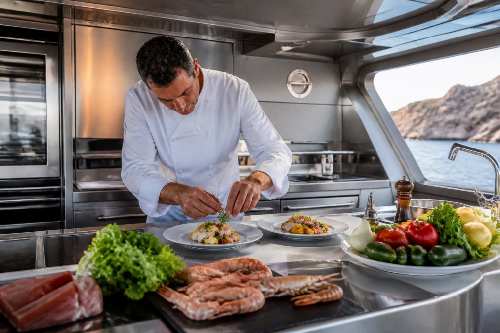 Professional onboard chef in white chef's jacket preparing gourmet Mediterranean cuisine in a modern, stainless-steel yacht galley, with fresh seafood, vegetables, and elegant plating visible on the counter