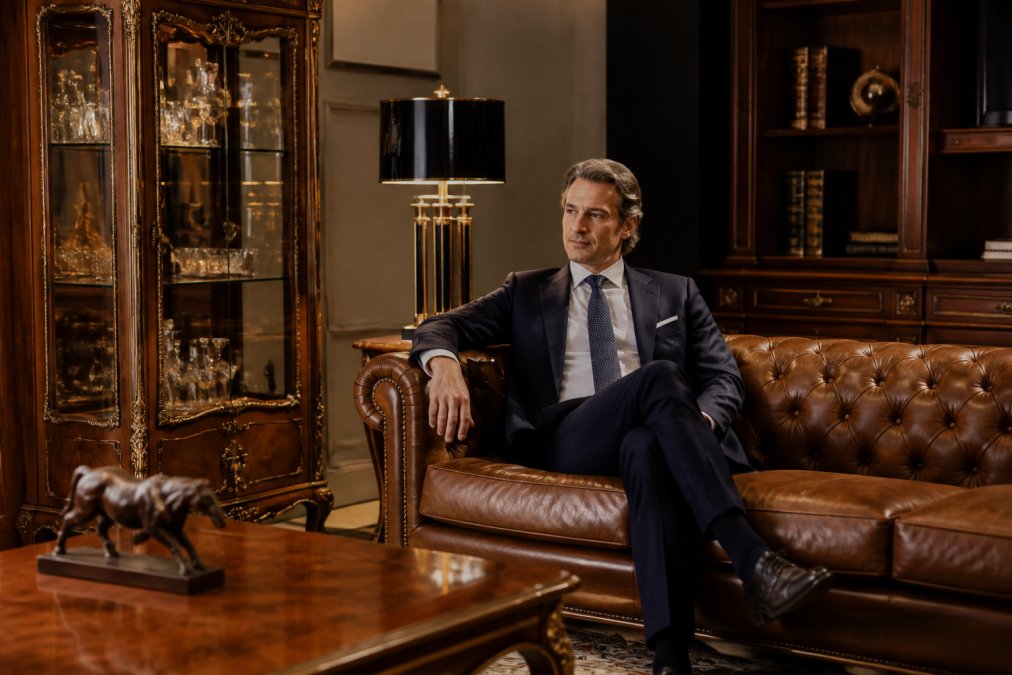 Ein luxuriöses Empfangsbüro mit Chesterfield-Sofa in Braun-Antik, handgeschnitzter Barock-Vitrine mit Glaseinsätzen, eleganter Tischleuchte im Art Deco Stil und hochwertigem Aktenschrank. Ein Geschäftspartner sitzt entspannt auf dem Sofa, die Raumgestaltung wirkt repräsentativ und vertrauenswürdig
