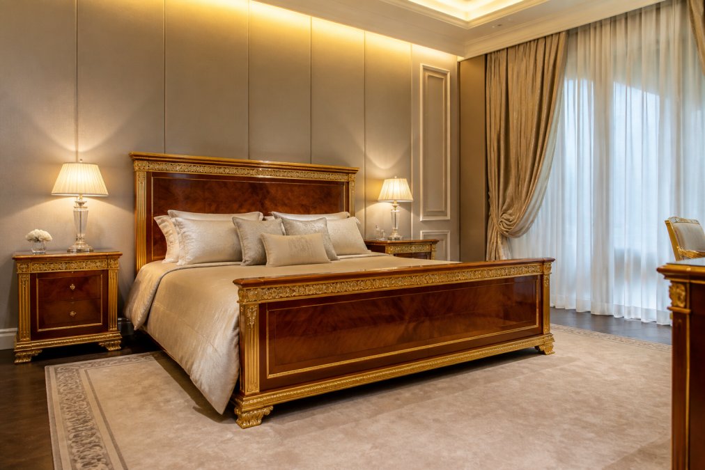 Luxuriöses Schlafzimmer einer Villa mit hochwertigem Massivholzbett mit Blattgold-Details, eleganten Nachttischen, klassischen Leuchtern und gedimmter indirekter Beleuchtung, Seidenvorhänge