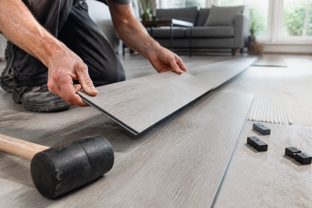 Close-up of hands snapping together two LVT planks with a click system in a bright, modern living room. The floor is partially installed, with a rubber mallet and spacers nearby.