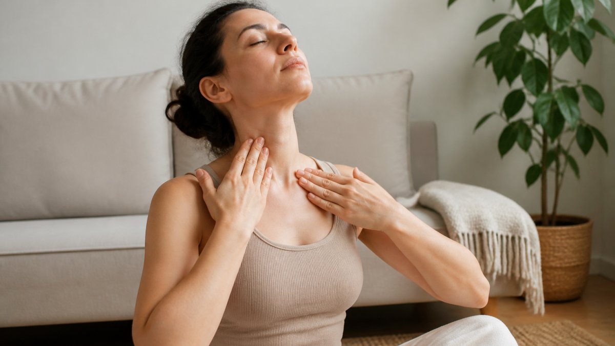 A woman in comfortable clothing gently massaging her neck and collarbone area with her fingertips in a calm, softly lit room at home