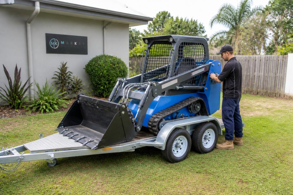 A compact mini loader on a trailer parked in a suburban backyard, with a 4-in-1 bucket attached and a person in work clothes checking the machine before use