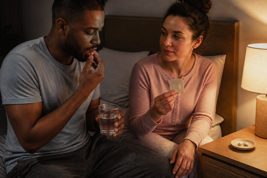 A bedside table at night with a glass of water, a small dish holding two capsules, and a sleep patch packet, soft lamp light in a calm bedroom