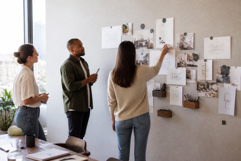 Modern open-plan office with team members collaborating at a wall covered in magnetic wallcovering, displaying project notes, sketches, and magnetic accessories under natural window lighting