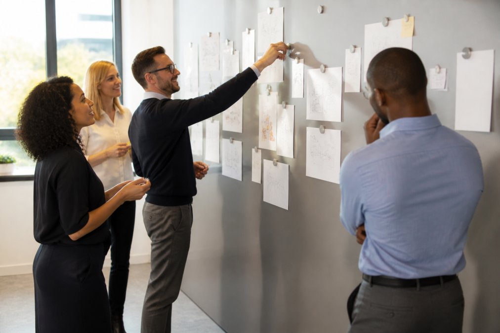 Diverse team of office workers actively collaborating around full magnetic wall in modern workspace, with notes, diagrams, and project sketches visible on the surface under natural office lighting