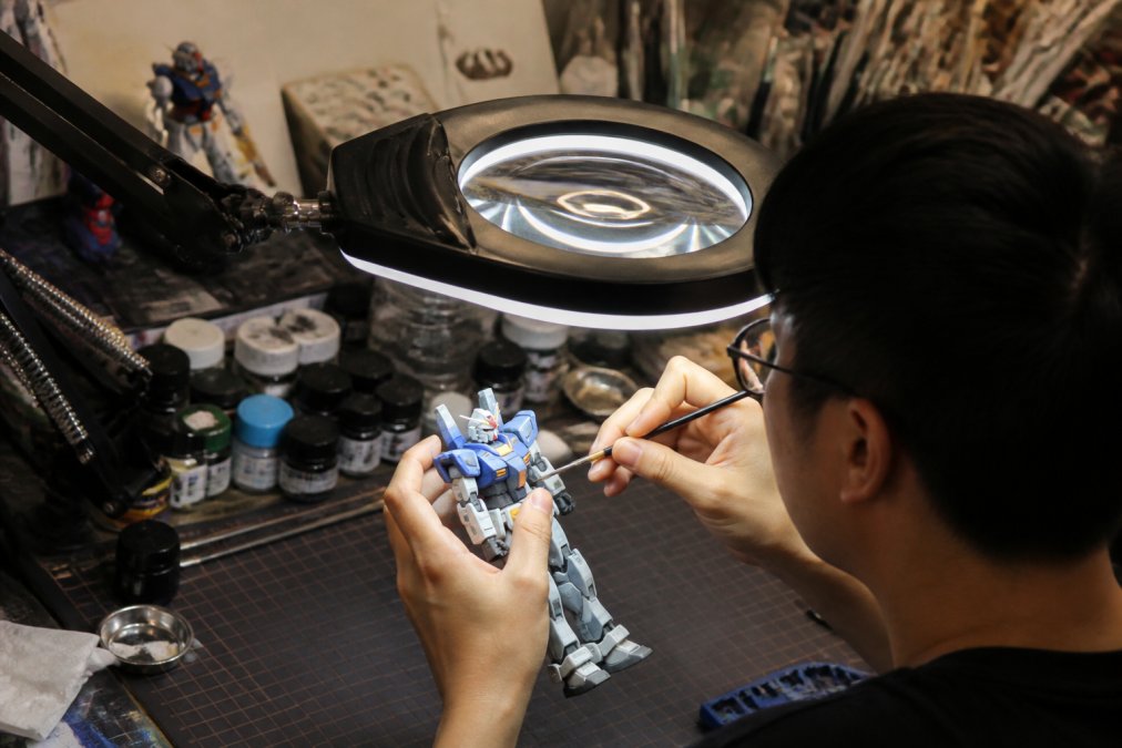 Close-up of a hobbyist's hands painting a small Gundam model kit under a bright LED magnifying lamp on a cluttered workbench, the shadow-free ring light illuminating the miniature while the lens stays in sharp focus