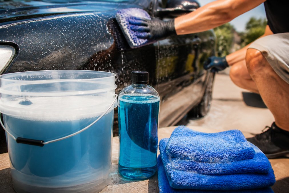 Hands washing a dark-colored car with microfiber wash mitt, applying pH-neutral shampoo under bright sunlight, water beading on the freshly washed surface