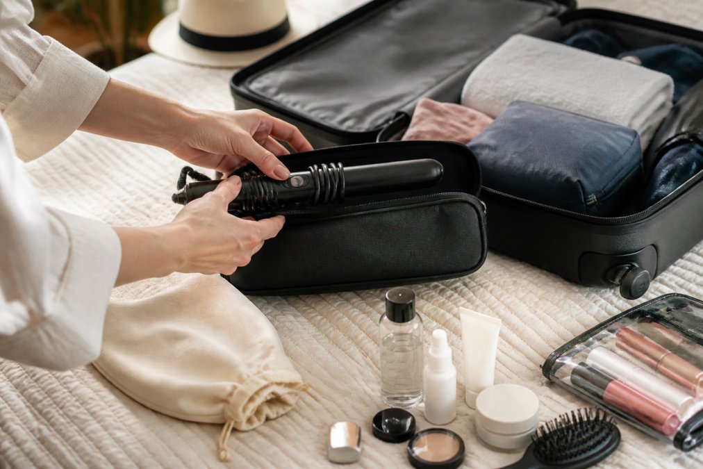 Woman packing a compact hair styling tool into a travel bag alongside toiletries and cosmetics, with natural daylight illuminating the organized luggage